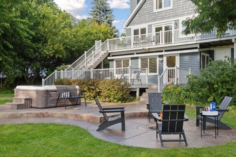 A backyard scene featuring a hot tub, fire pit, and Adirondack chairs in front of a two-story house with a deck.