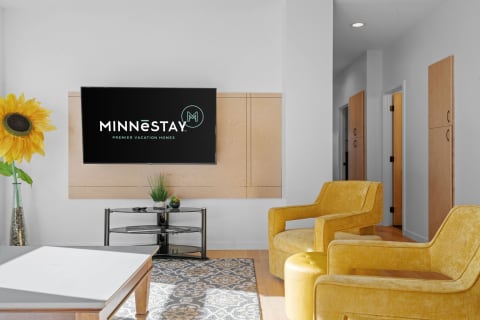 Stylish living room with a MINNēSTAY logo on a television, yellow chairs, and a sunflower decoration.
