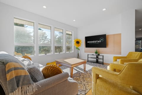A modern living room with large windows, a gray sofa, yellow chairs, and a TV.