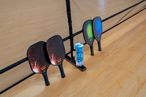 Two pairs of pickleball paddles and a holder with blue balls near a net on wooden flooring.