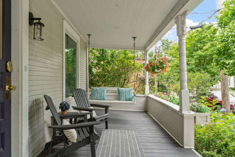 A front porch featuring dark wooden chairs, a hanging swing with turquoise pillows, and vibrant plants.