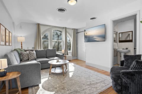 A bright living area with a gray sectional sofa, wooden coffee table, and plush armchair, illuminated by large arched windows.