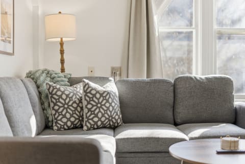 A gray sectional sofa with patterned pillows and a cozy throw, with natural light coming through windows.