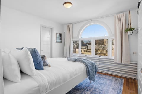 A bright bedroom featuring a bed with white linens and blue accents, arched windows with natural light, and wooden flooring.