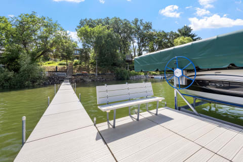 A wooden dock with a white bench and a boat on calm water surrounded by trees.