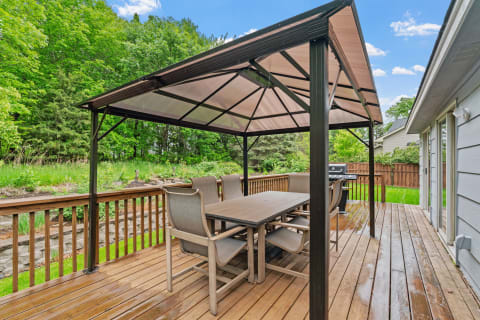 Outdoor dining space with a gazebo and plush chairs on a wooden deck surrounded by trees.