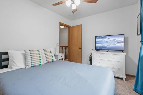 A cozy bedroom featuring a blue duvet, colorful striped pillows, a white dresser with a TV, and a wooden door.