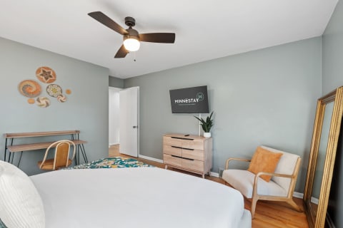 Cozy modern bedroom with a desk, dresser, and wall art, featuring a bed and armchair.