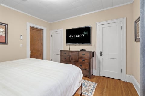 A cozy bedroom with a bed, wooden dresser, and mounted TV displaying MINNESTAY logo.