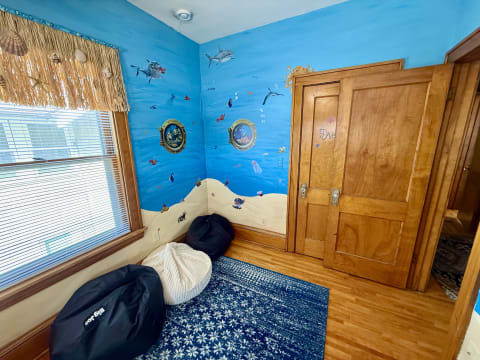A children's room with blue walls featuring sea life murals, bean bags, and a window with a straw valance.