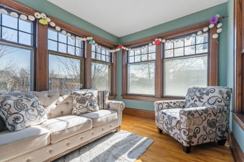 A bright sunroom featuring a tufted sofa, patterned armchair, and colorful window decorations.