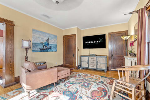 Interior view of a living room with brown sofa, colorful rug, painting of a boat, and a TV on the wall.