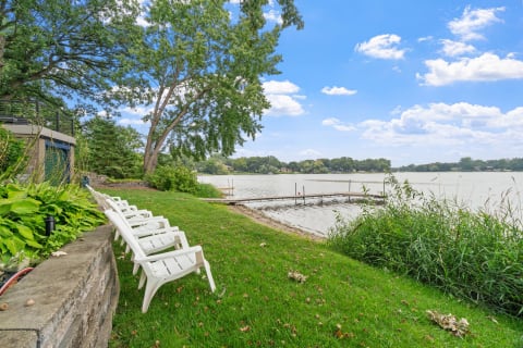 Lakeside scene featuring white chairs, greenery, and a wooden dock.