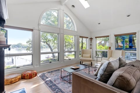 Modern living room with large arched windows and lake view, featuring beige armchairs and decorative poufs.