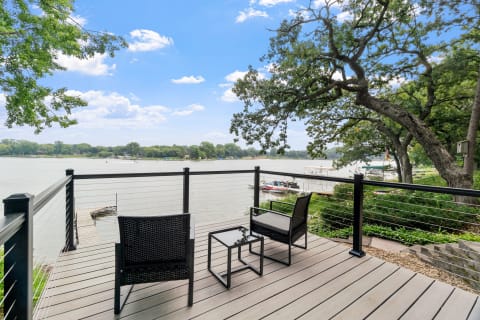 Outdoor deck with chairs and a table beside a calm water body, featuring greenery and a dock.