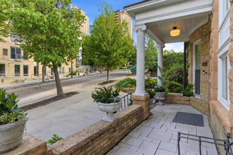 A charming property entrance featuring white columns and potted plants, with a quiet street and modern buildings in the background.