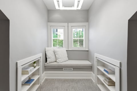 A small reading nook with a cushioned bench, plantation shutters, and white shelves filled with books.