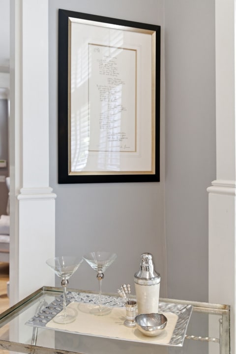An elegant bar setup with martini glasses, a cocktail shaker, and stirring sticks on a shiny tray against a gray wall.