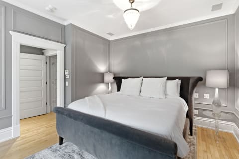 A stylish bedroom with plush white bedding, gray velvet headboard, and warm wood floors, illuminated by ambient lighting.