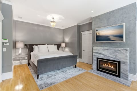 A cozy and elegant bedroom with gray walls, a plush bed, a modern fireplace, and a beautiful landscape photograph.