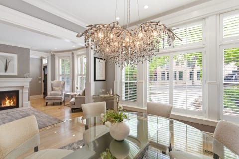 Elegant dining room with a glass table, plush chairs, and a stunning crystal chandelier, featuring a cozy fireplace and large windows.