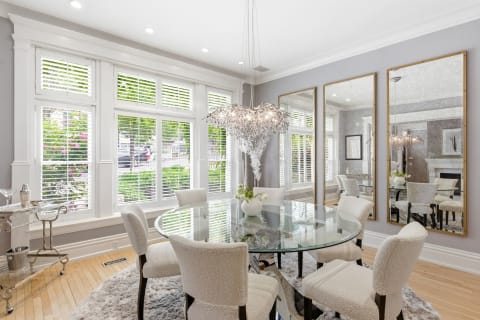 A bright dining room with a glass table, soft chairs, and a unique chandelier, surrounded by large windows.