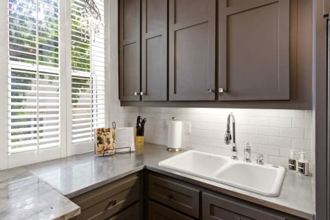 A modern kitchen with dark cabinets, marble countertops, and a farmhouse sink under natural light from plantation shutters.