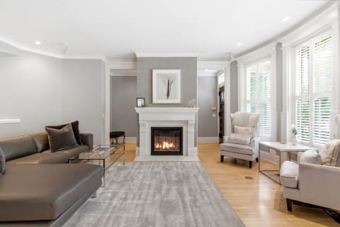 Modern living room with a fireplace, leather sofa, and plush armchairs, brightened by natural light from large windows.
