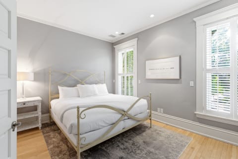 A cozy bedroom with a decorative bed, soft linens, gray walls, and light streaming in through shutters.