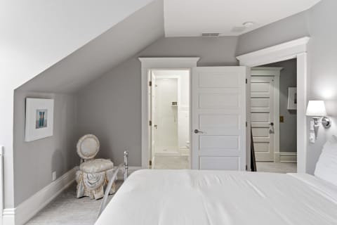 A cozy bedroom suite with soft gray walls and an elegant stool, showcasing a door to a bright ensuite bathroom.