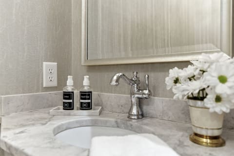 A marble bathroom countertop with a vintage chrome faucet, bottles of lotion and hand soap, and white flowers in a gold-trimmed pot.