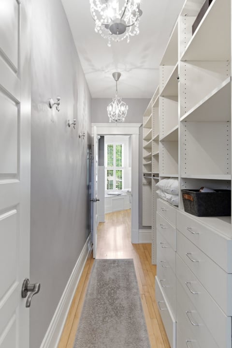 A spacious walk-in closet with elegant chandeliers and organized white shelving, leading to a bright window nook.