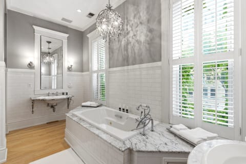Elegant bathroom featuring a marble soaking tub, crystal chandelier, and natural light with plantation shutters.