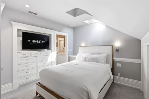 A modern bedroom featuring a large bed with white bedding, a mounted TV, and soft gray walls, illuminated by natural light and stylish sconces.