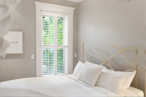 A cozy bedroom featuring a white iron bed, crisp linens, and a window with plantation shutters letting in natural light.