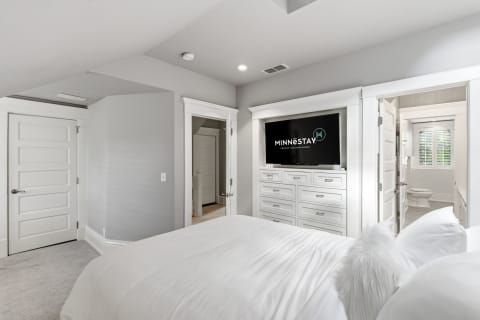 A tranquil bedroom featuring a white bed, modern dresser with TV, and access to a private en-suite bathroom.