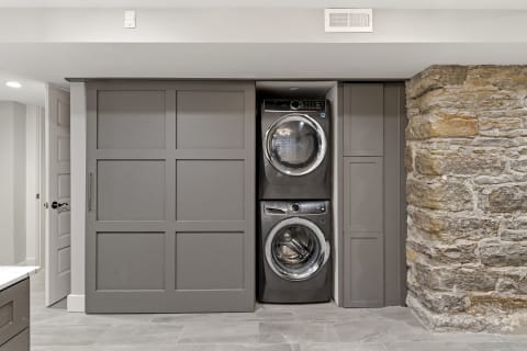 Modern laundry space featuring stacked washer and dryer behind gray cabinetry and a stone wall.