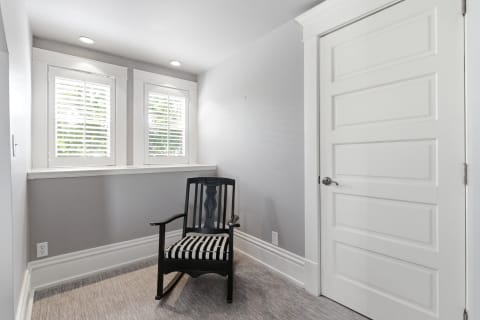 A black rocking chair with a striped cushion in a bright, minimalist reading nook with gray walls and white shutters.