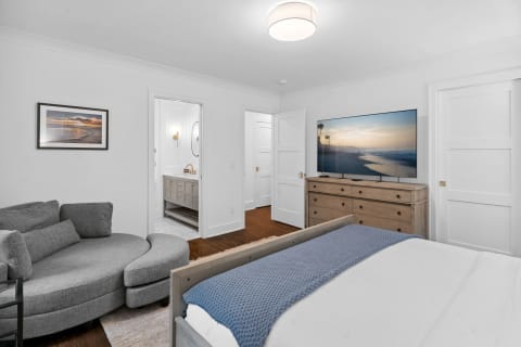 A modern bedroom featuring a gray sofa, white bed, and TV displaying a beach scene.