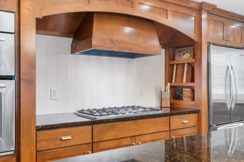 A modern kitchen with a gas cooktop under a wooden hood and dark granite countertops.