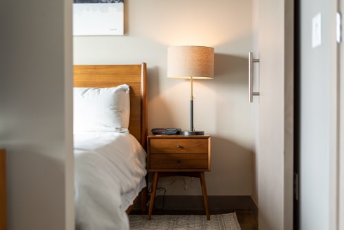 Sink into crisp white linens beside your stylish wooden nightstand, where warm lamplight creates the perfect reading nook for peaceful evenings.