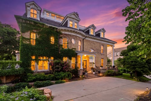 Ivy-draped Victorian elegance welcomes arrivals beneath a stunning purple-pink sunset. The historic brick facade glows with warm interior lighting, framed by mature gardens and a welcoming entrance.