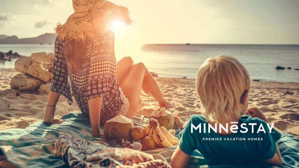 Woman on the beach of Lake Superior on a blanket with a child, sun setting in the distance. Minnestay logo bottom right
